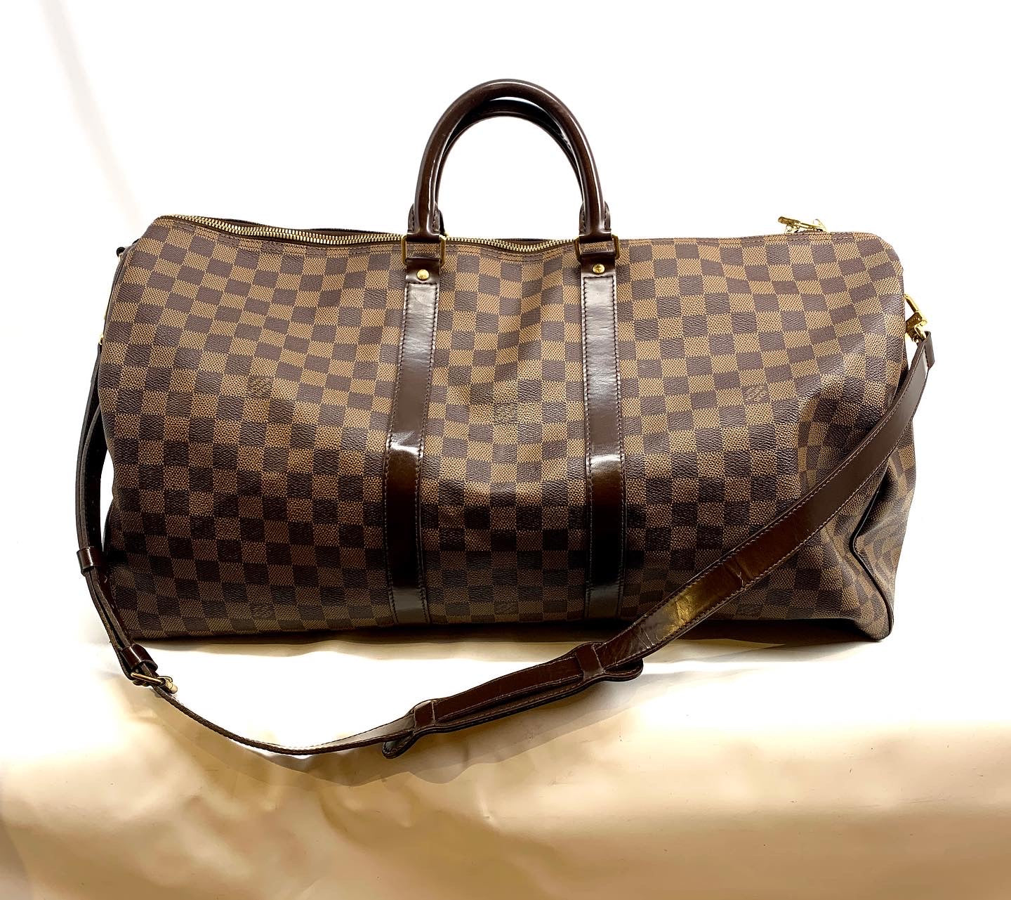 A Louis Vuitton monogram duffle bag in brown and beige checkered canvas with dark brown leather handles and trim, shown on a light beige fabric surface.
