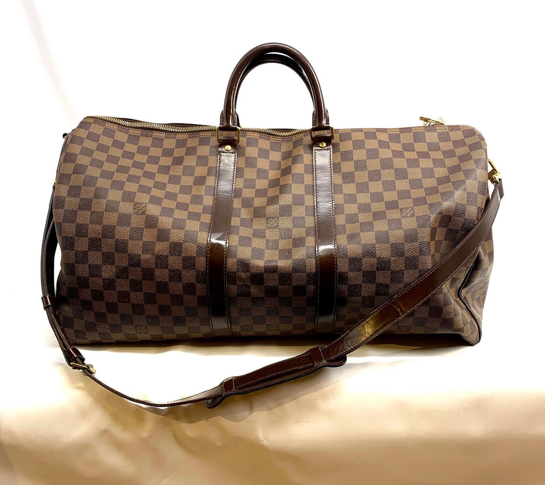 A Louis Vuitton monogram duffle bag in brown and beige checkered canvas with dark brown leather handles and trim, shown on a light beige fabric surface.