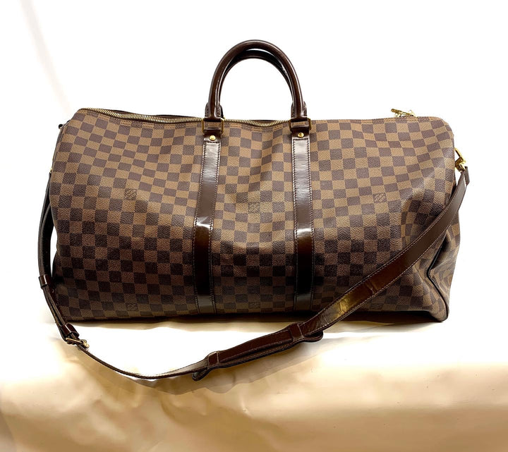 A Louis Vuitton monogram duffle bag in brown and beige checkered canvas with dark brown leather handles and trim, shown on a light beige fabric surface.